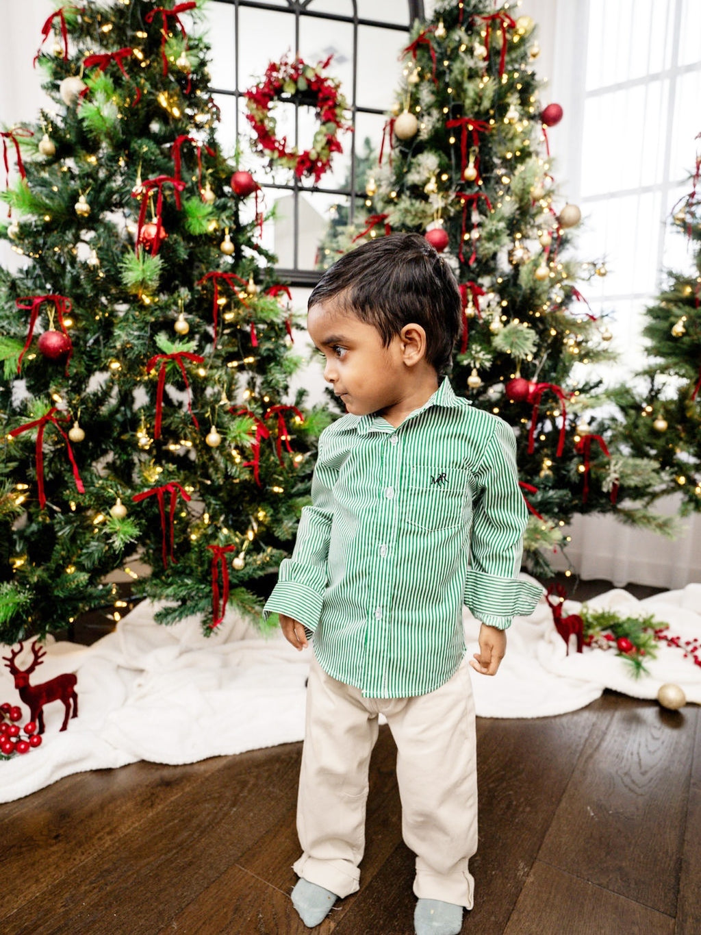 front full shot of a toddler boy wearing a white and green shirt with MAR embroidery on the pocket with white/cream pants in front of Christmas trees