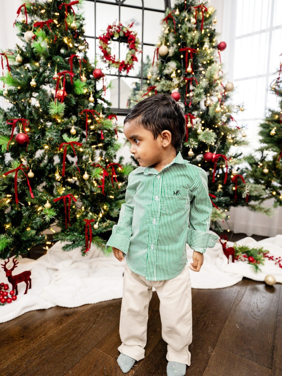 front full shot of a toddler boy wearing a white and green shirt with MAR embroidery on the pocket with white/cream pants in front of Christmas trees