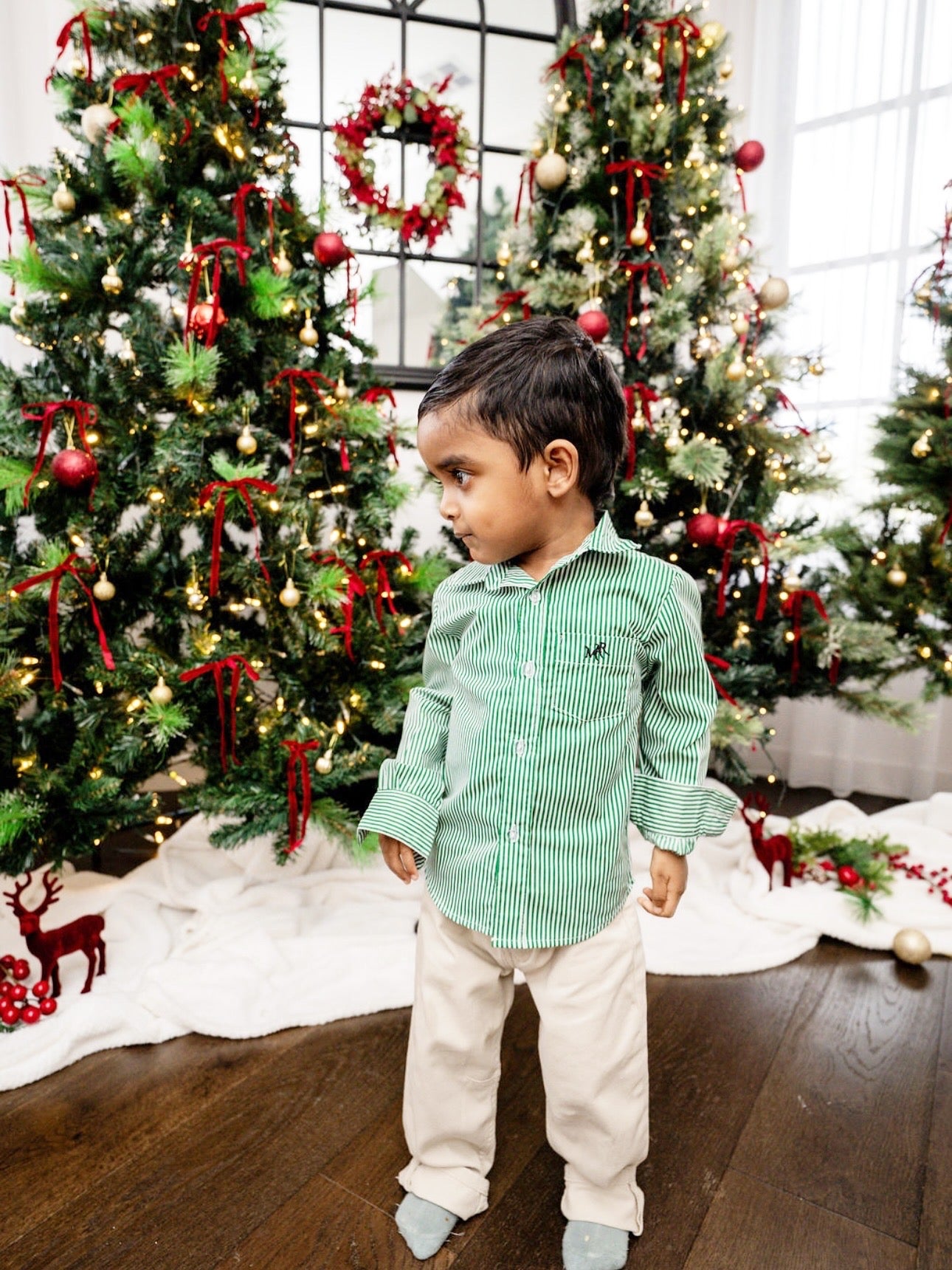 front full shot of a toddler boy wearing a white and green shirt with MAR embroidery on the pocket with white/cream pants in front of Christmas trees