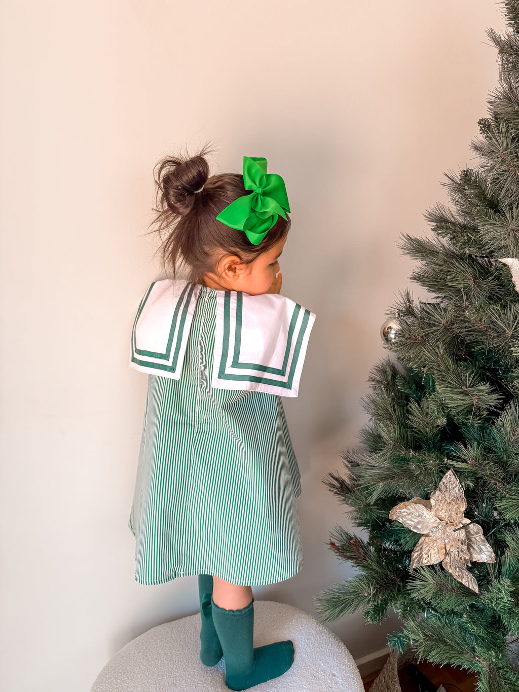 Child in a green and white dress with a large green bow standing next to a decorated Christmas tree.