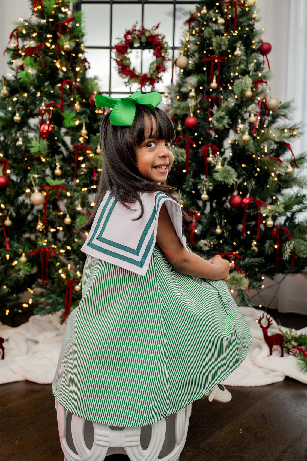 Child in a green dress with a bow standing in front of Christmas trees.