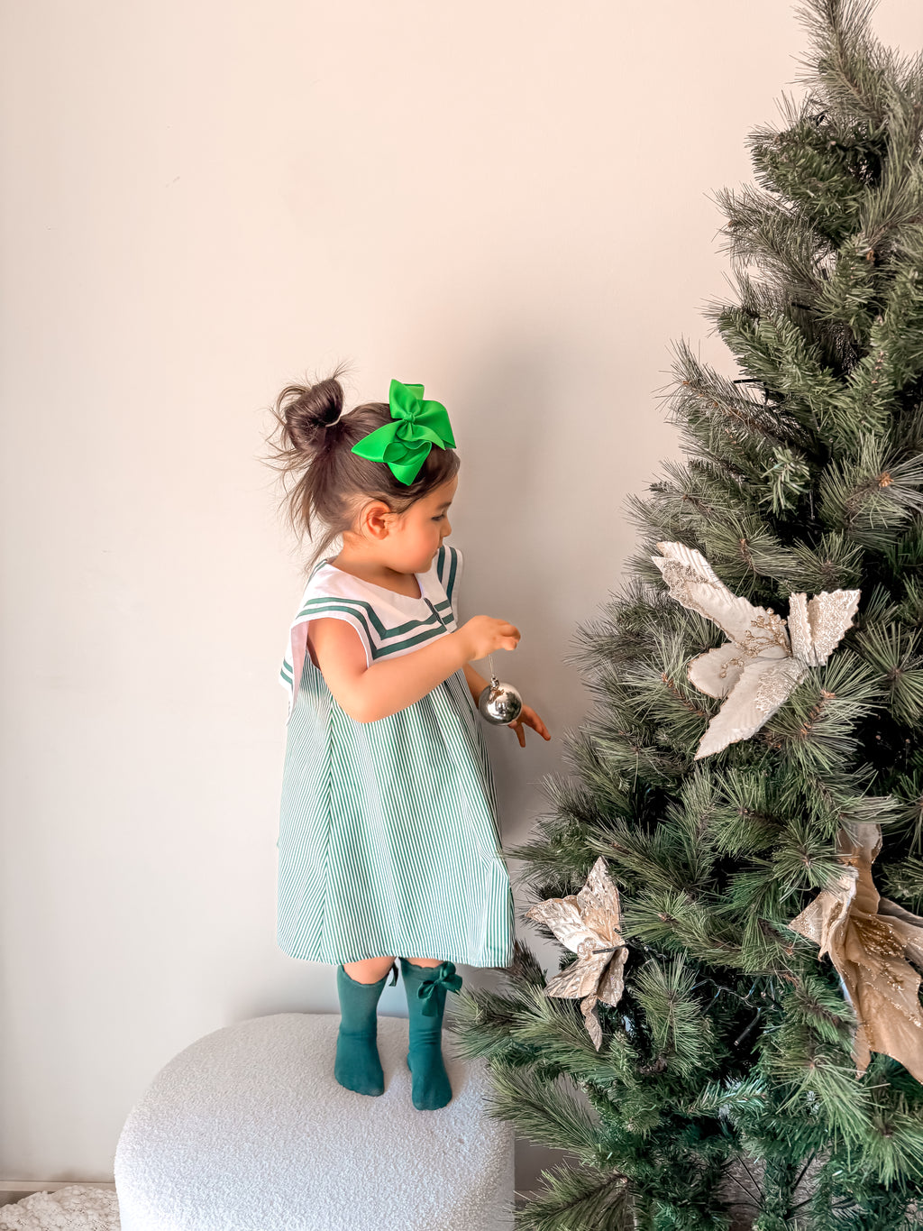 Child in a light blue dress with a green bow standing next to a decorated Christmas tree.