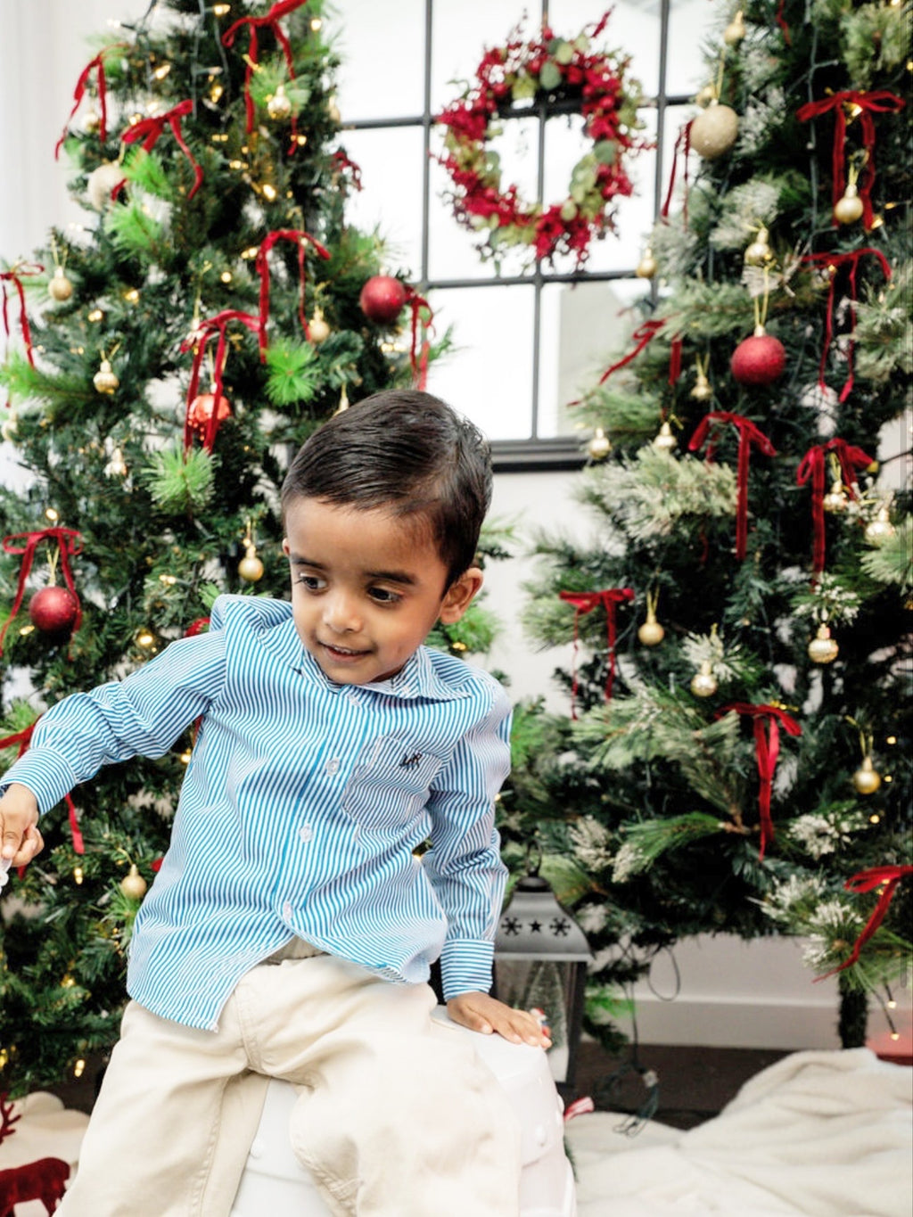 front full shot of a toddler boy wearing a white and blue shirt with MAR embroidery on the pocket with white/cream pants in front of Christmas trees