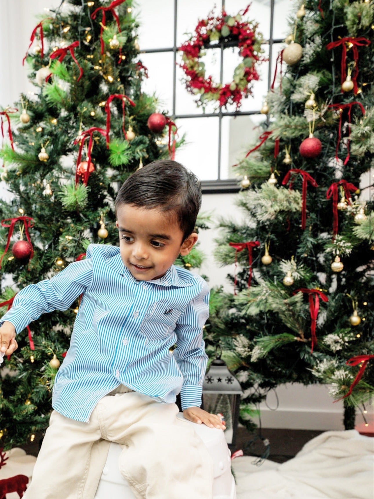 front full shot of a toddler boy wearing a white and blue shirt with MAR embroidery on the pocket with white/cream pants in front of Christmas trees