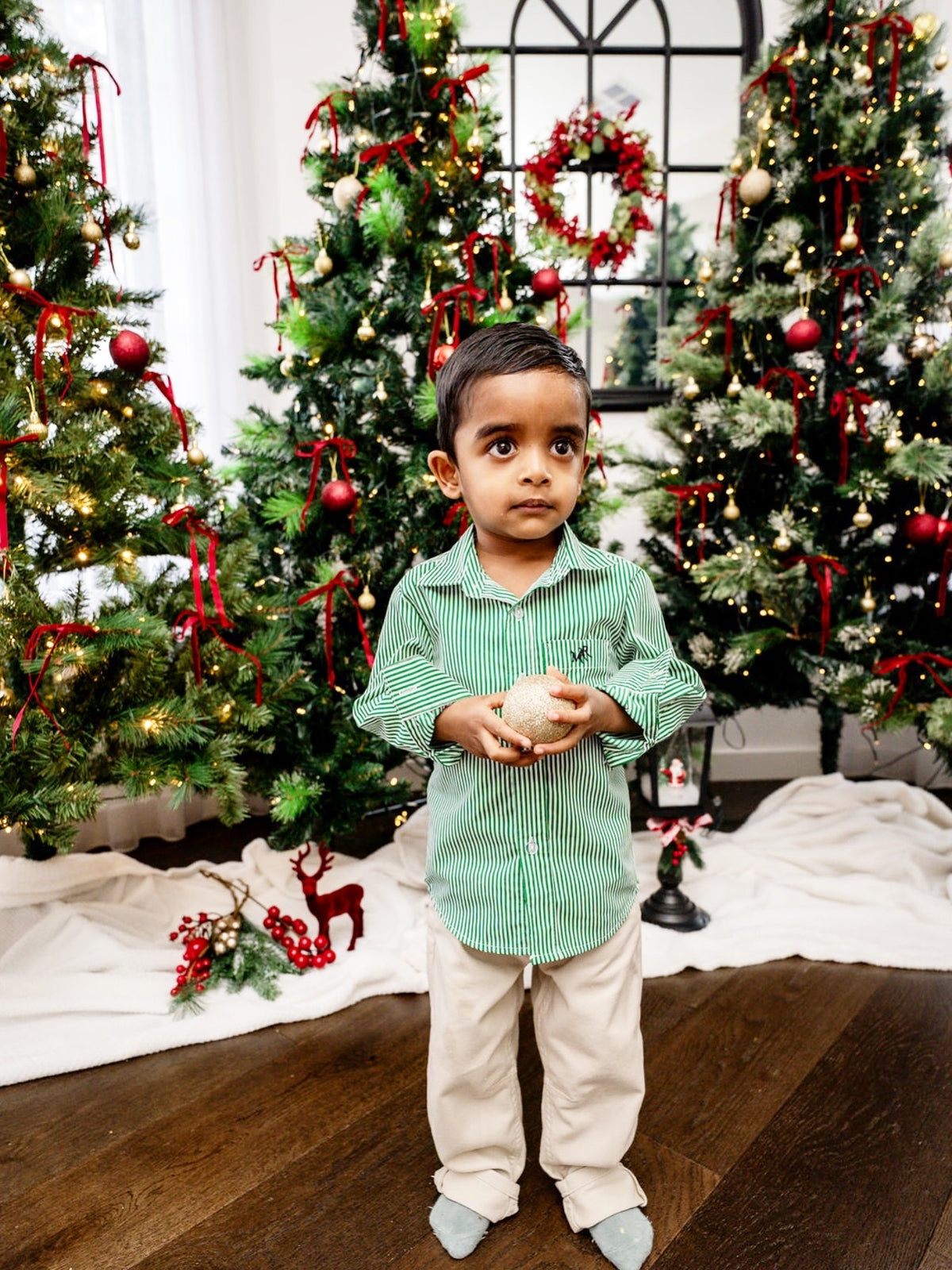 front profile photo- full shot of a toddler boy wearing a white and green shirt with MAR embroidery on the pocket with white/cream pants in front of Christmas trees