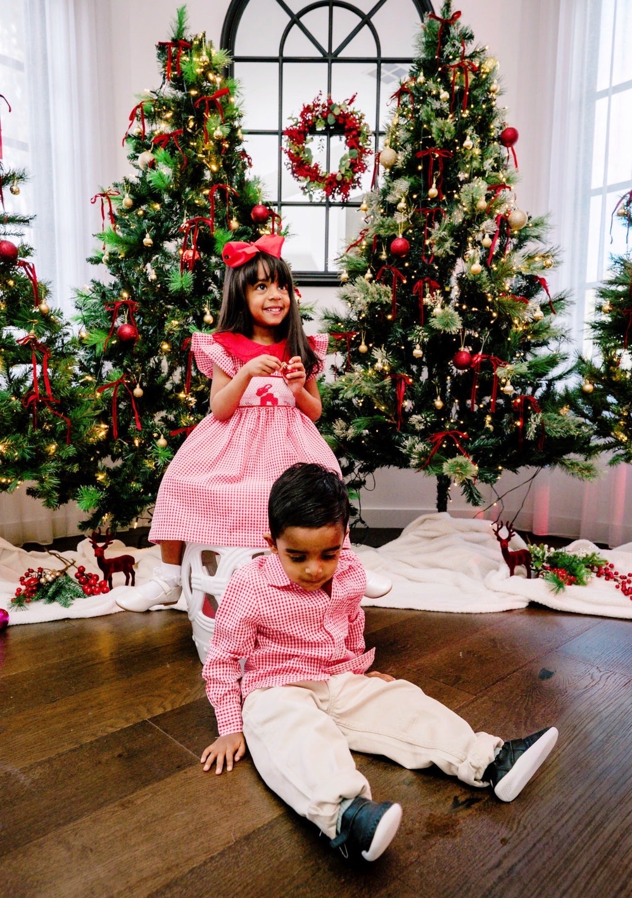 Two children in festive attire sitting on a wooden floor with decorated Christmas trees in the background.