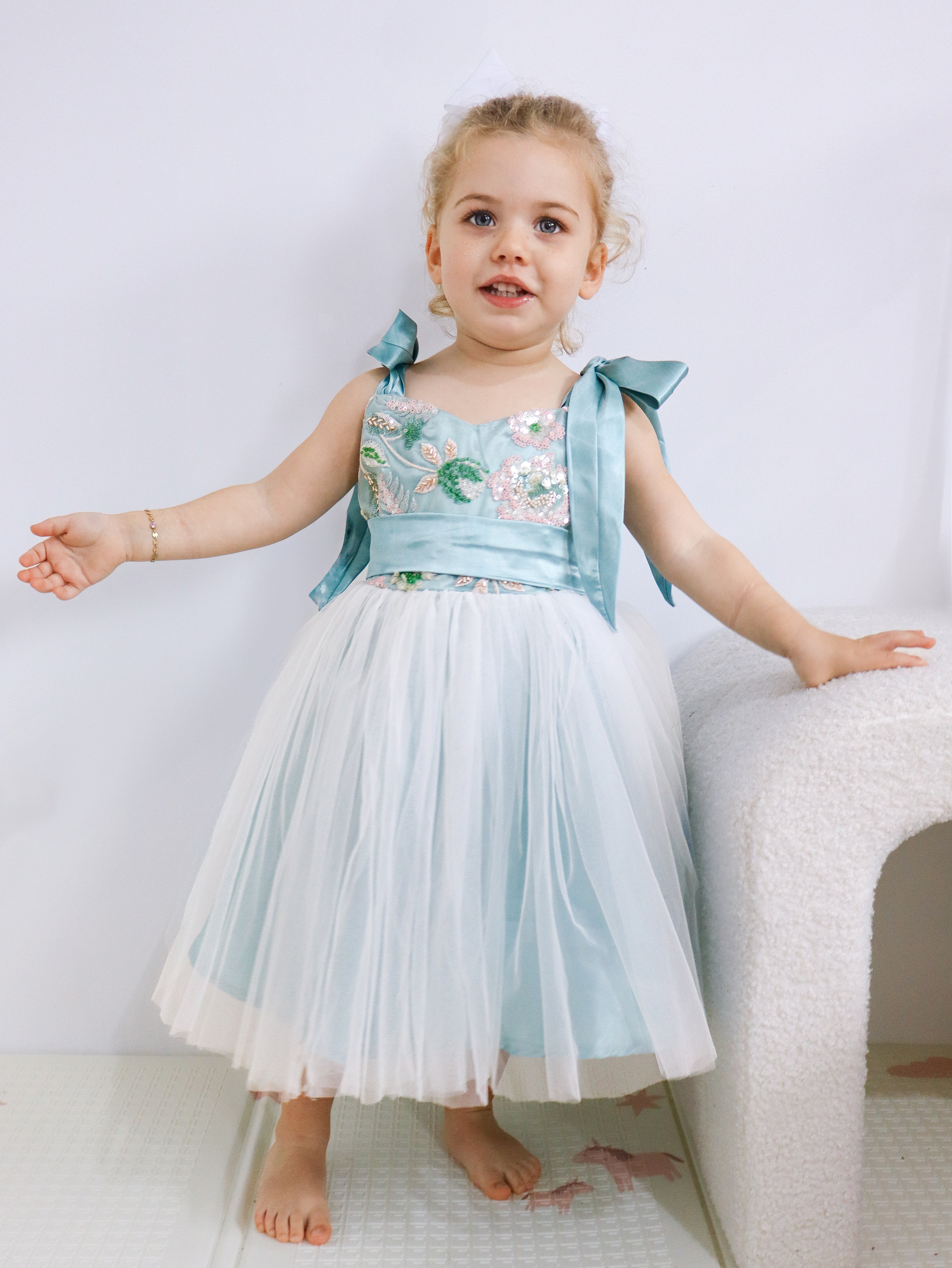 Young girl in a light blue and white dress with floral embroidery, standing on a white surface.