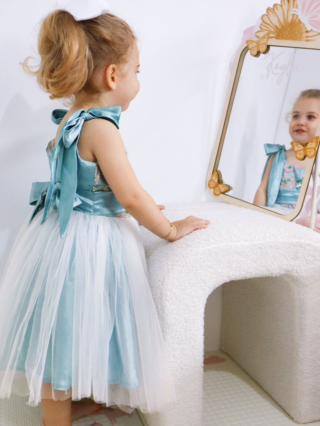 Child in a light blue and white dress looking into a mirror.