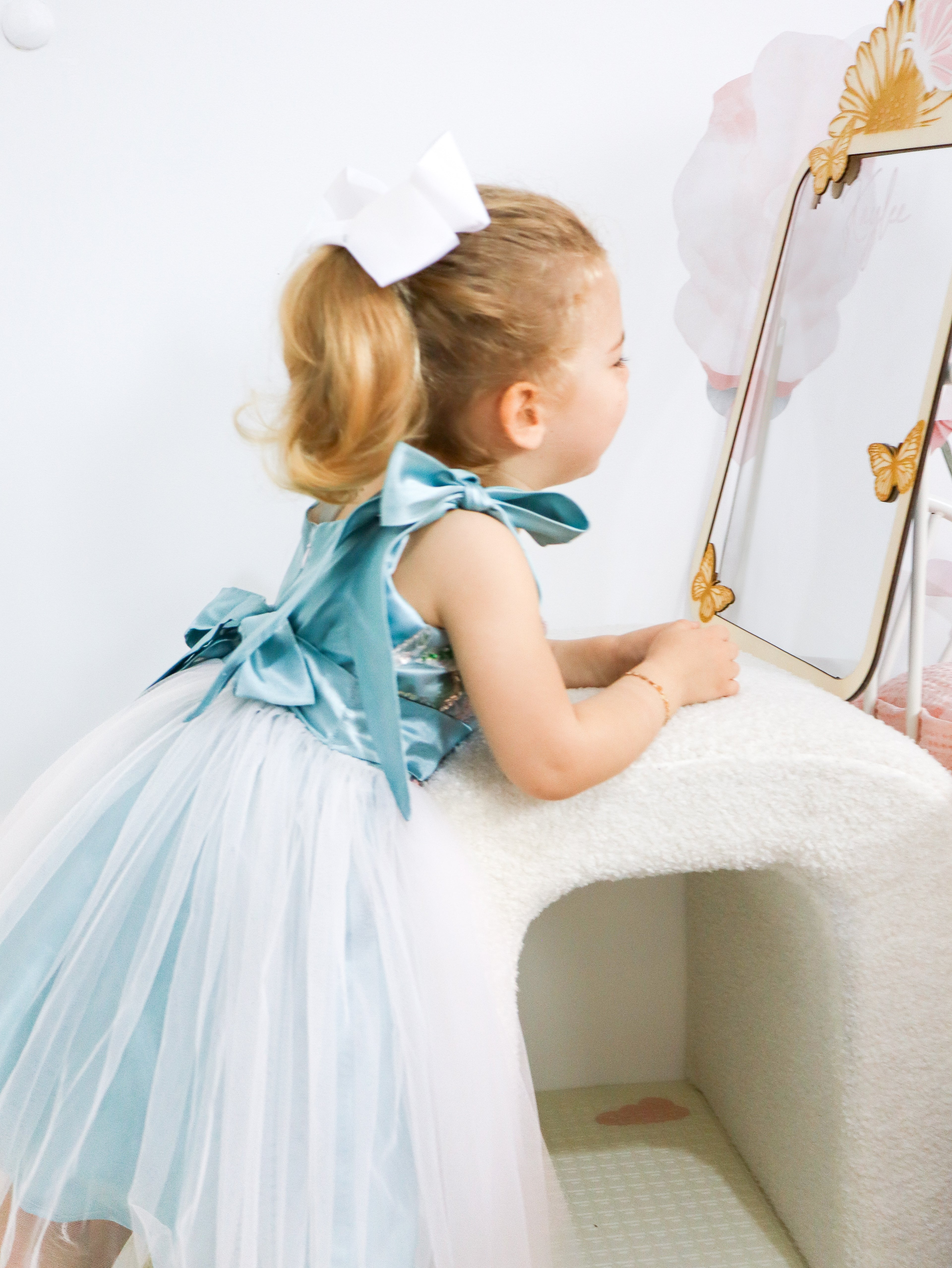 Young girl in a light blue dress with a large bow sitting on a white surface.