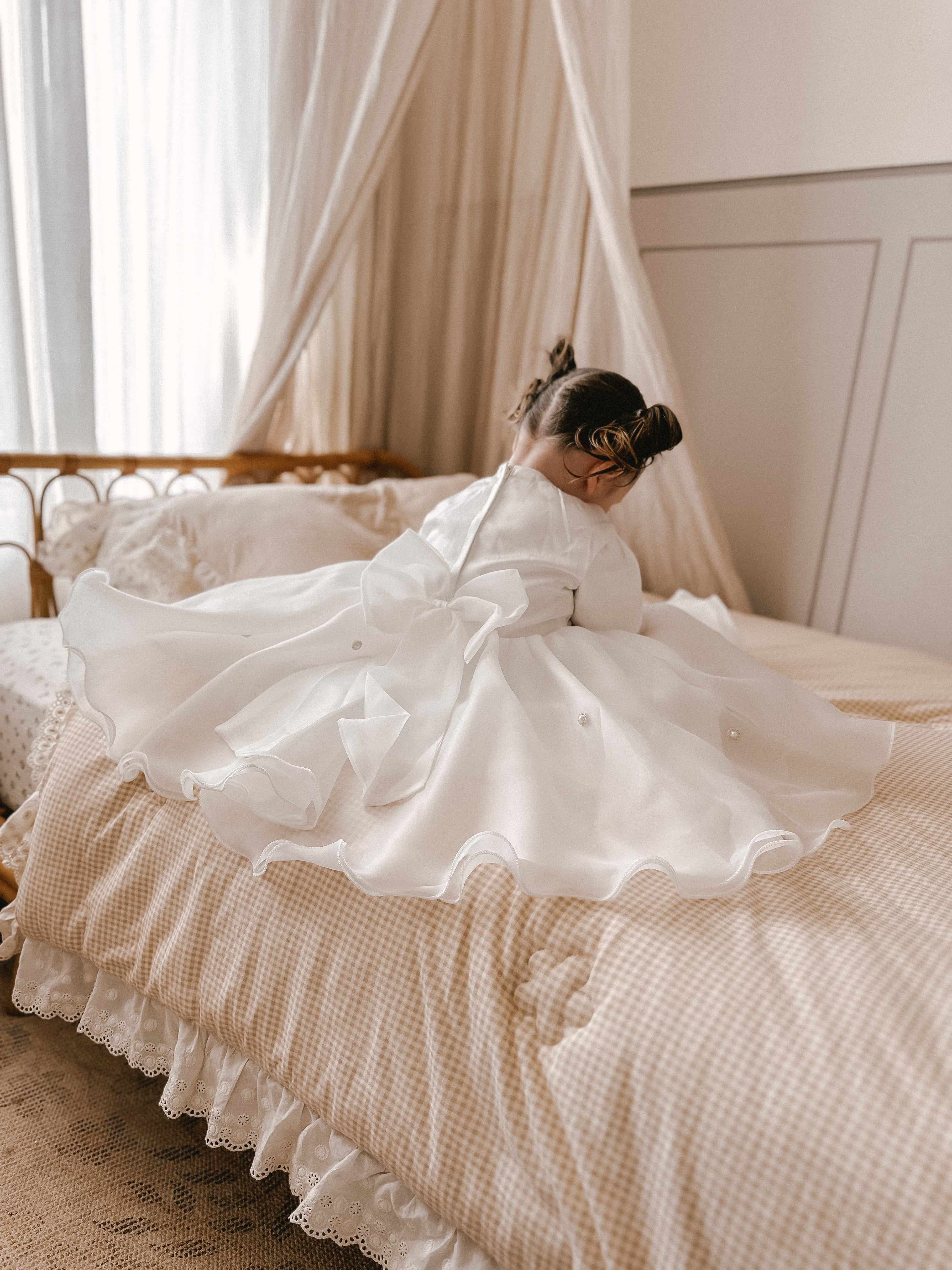 Child in a white dress sitting on a bed with a canopy in a softly lit room wearing a white satin dress with pearl embellishments. back of the dress is shown here which is beautiful and flowy nicely spread out on the bed with the big bow at the back. 