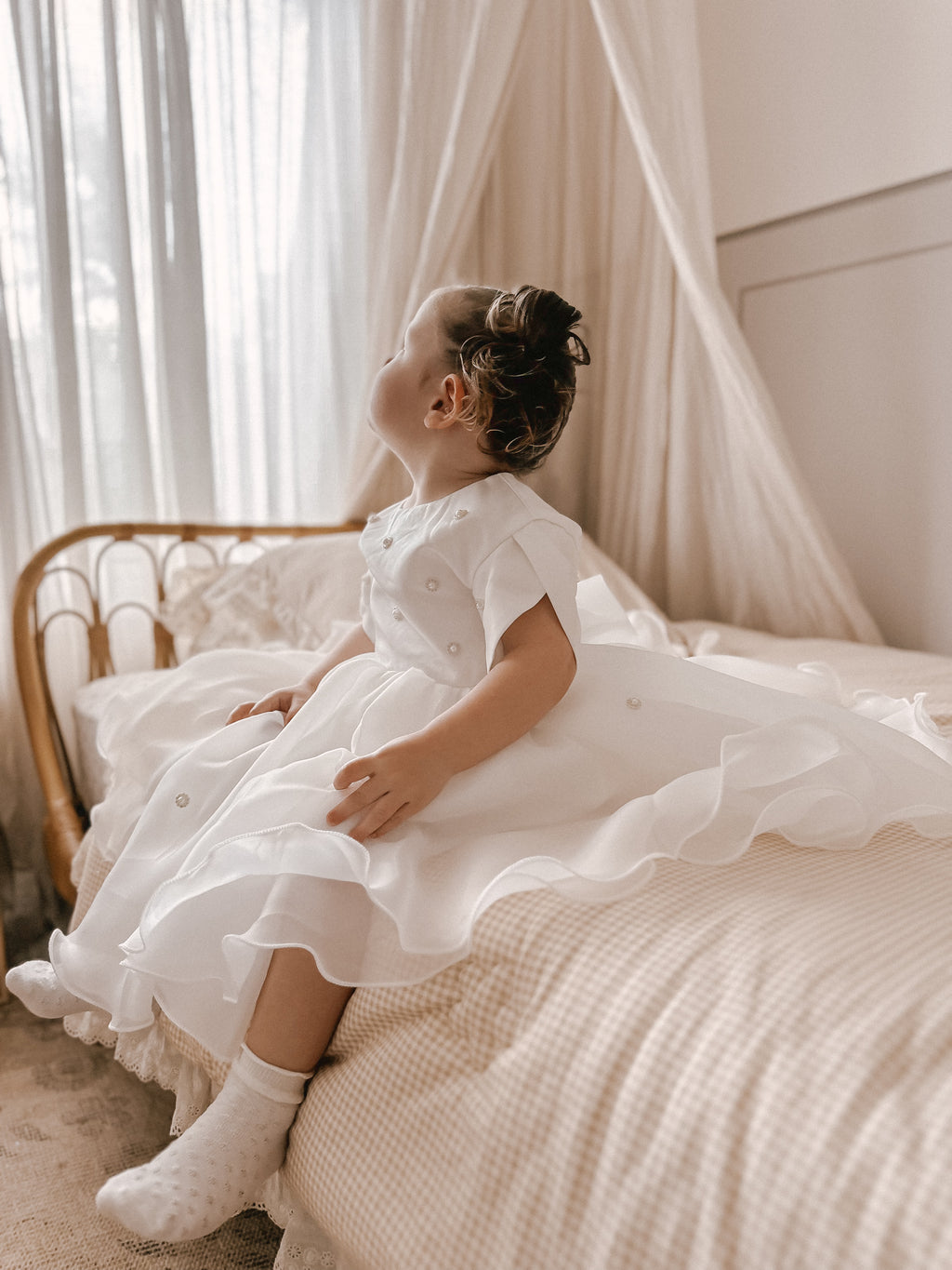 Young girl in a white dress sitting on a bed with white curtains in the background