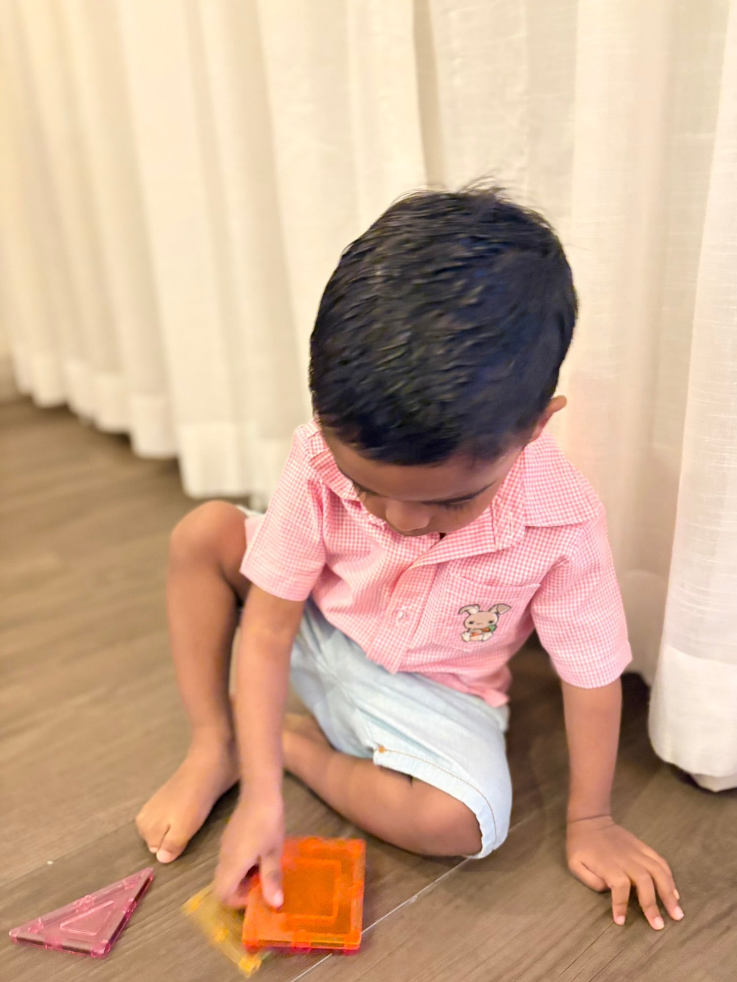 Child playing with colorful blocks on a wooden floor wearing a pink and white gingham short sleeve shirt made of 100% cotton fabric and it has a small pocket on the left side with a embroidered bunny. 