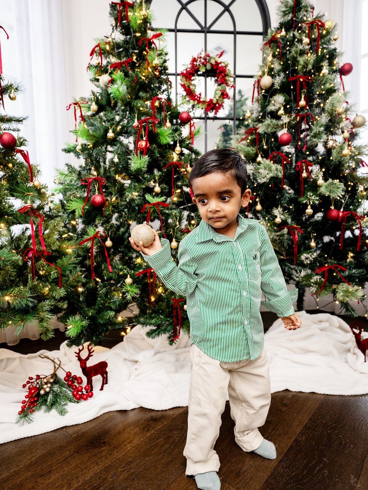 side shot of a toddler boy wearing a white and green shirt with MAR embroidery on the pocket with white/cream pants in front of Christmas trees