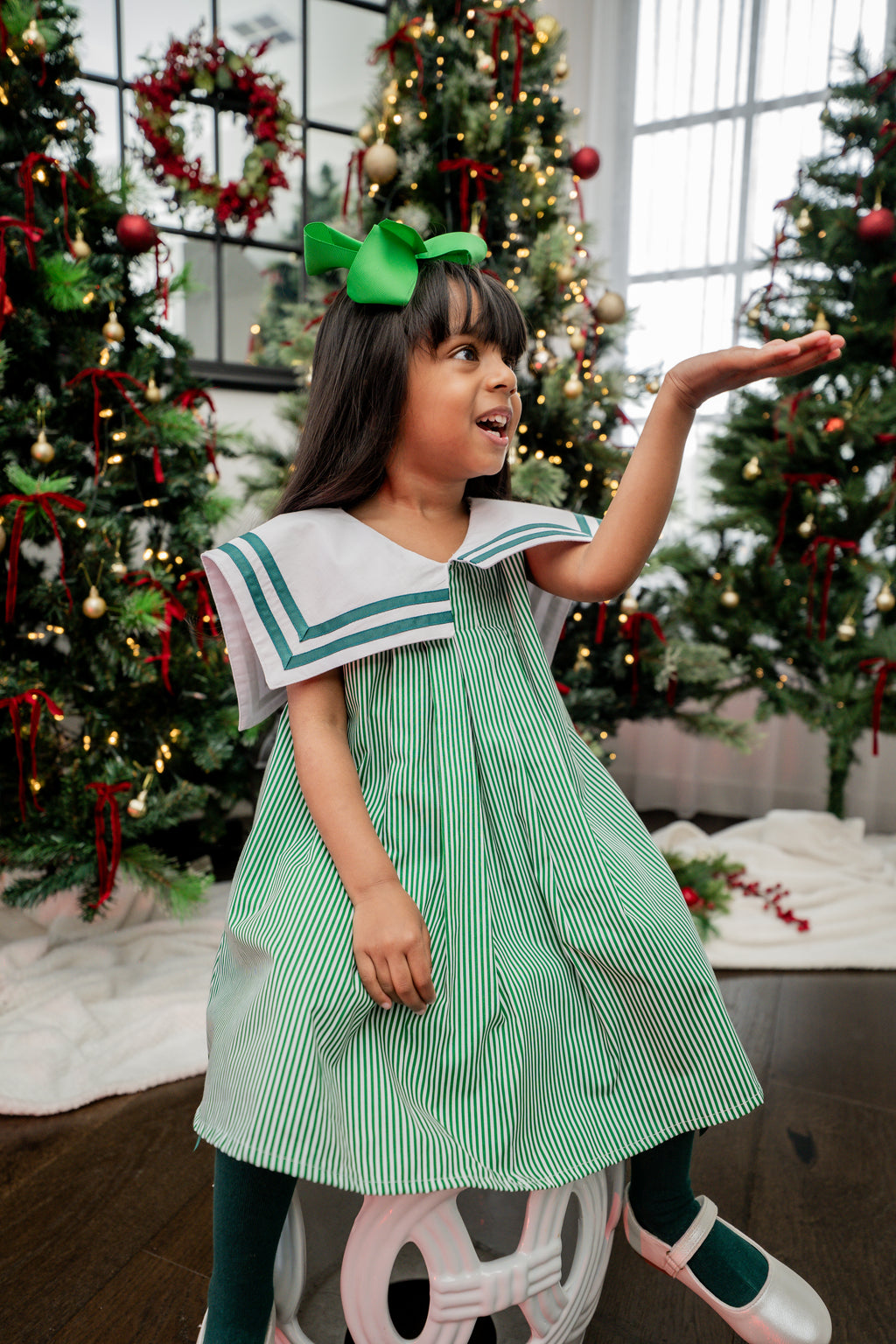 Young girl in a green dress with a white collar standing in front of Christmas trees.