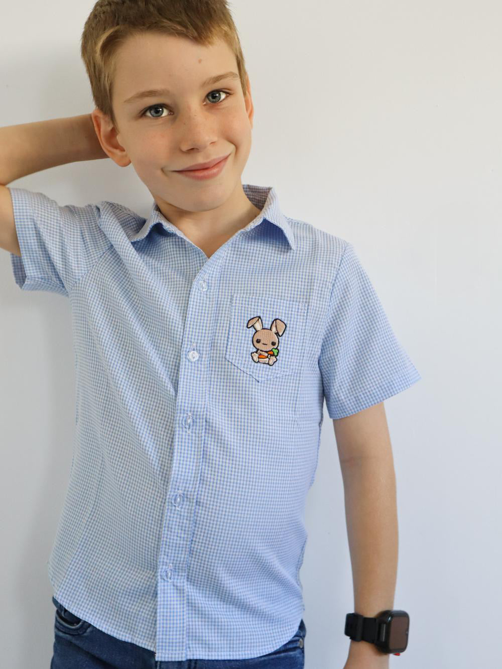 standing- boy in a white and blue gingham shirt made of cotton with a cute embroidered bunny on the side pocket. short sleeve
